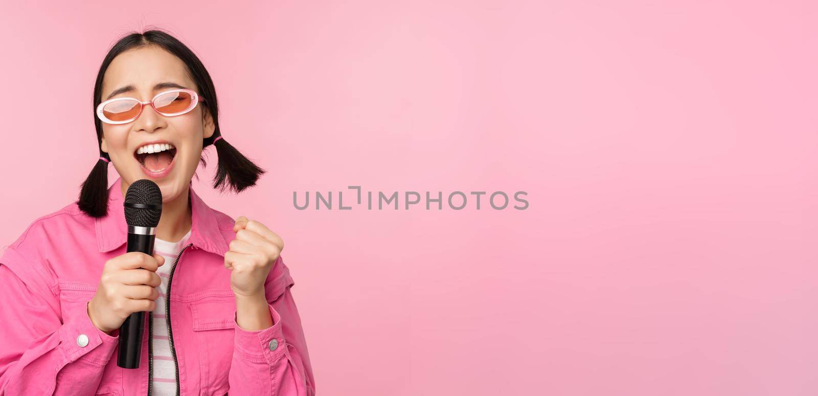 Happy beautiful asian girl singing with mic, using microphone, enjoying karaoke, posing against pink studio background by Benzoix