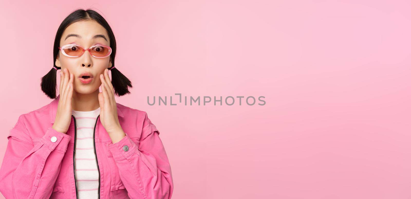 Image of asian girl looking surprised and excited, smiling, amazed reaction to big news, standing over pink background.