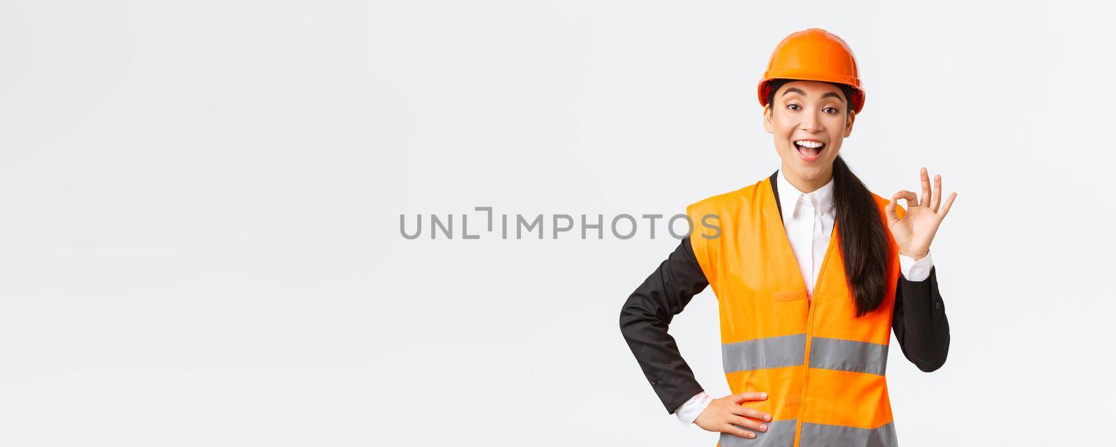 Satisfied happy asian female engineer, construction manager in safety helmet and reflective jacket showing okay gesture pleased, guarantee building quality, ensure everything excellent.