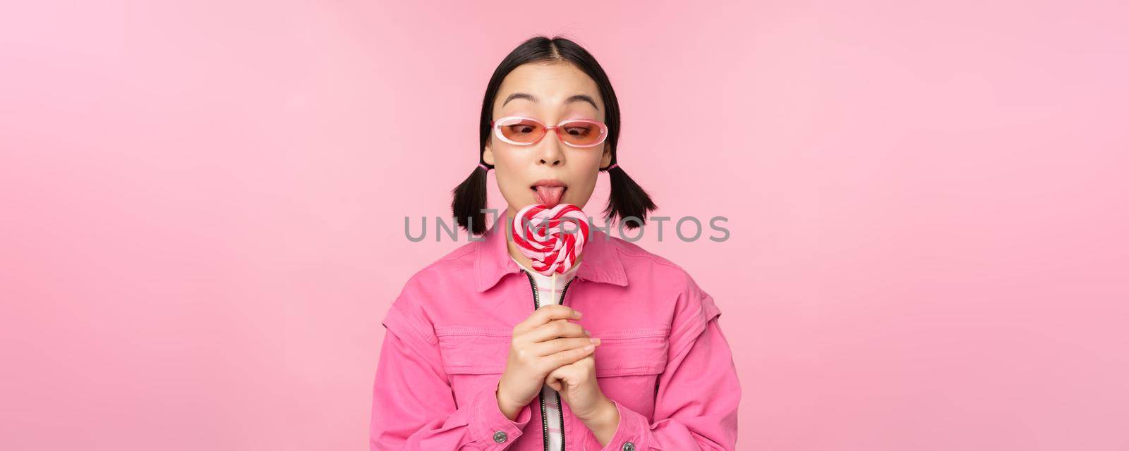 Silly and cute asian female model licking lolipop, eating candy sweet and smiling, looking excited, standing over pink background by Benzoix