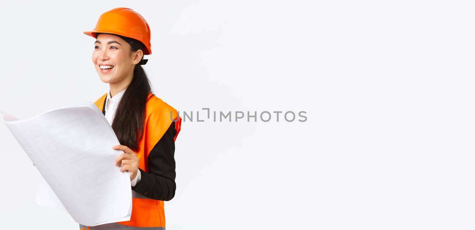 Satisfied happy asian female chief engineer looking at results of construction works, studying blueprint with pleased face, wearing safety helmet and reflective clothings at building zone.