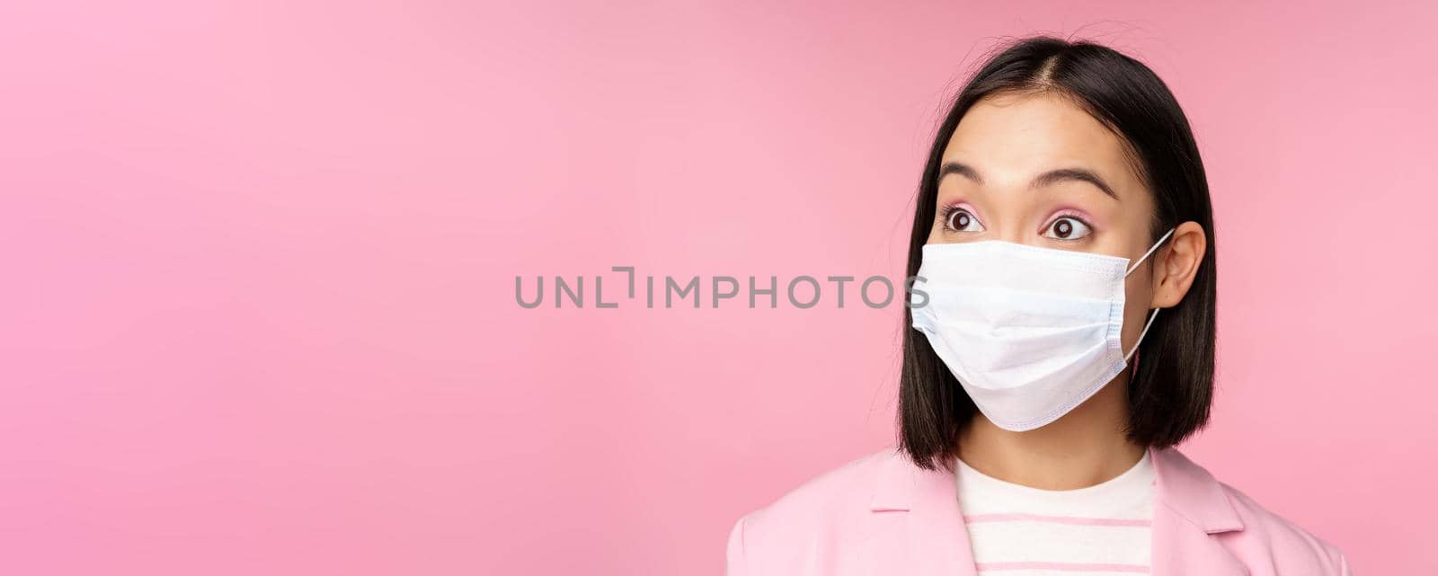 Close-up portrait of asian businesswoman in medical face mask, looking surprised, standing in suit over pink background. Copy space