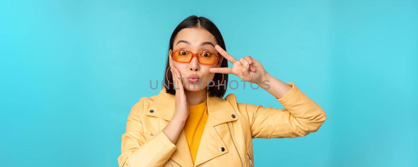 Portrait of attractive korean woman in sunglasses, showing peace v-sign near eyes, pucker lips, kissing gesture, standing over blue background.