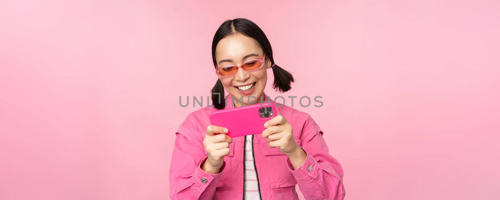 Portrait of happy asian girl playing on smartphone, watching videos on mobile phone app, standing over pink background.