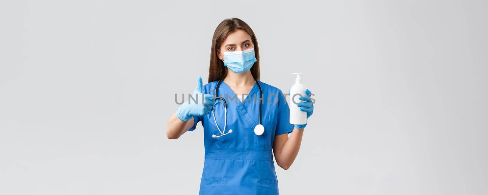 Covid-19, preventing virus, health, healthcare workers and quarantine concept. Confident female doctor, nurse in blue scrubs in medical mask and scrubs advice using hand sanitizer, thumbs-up approval by Benzoix