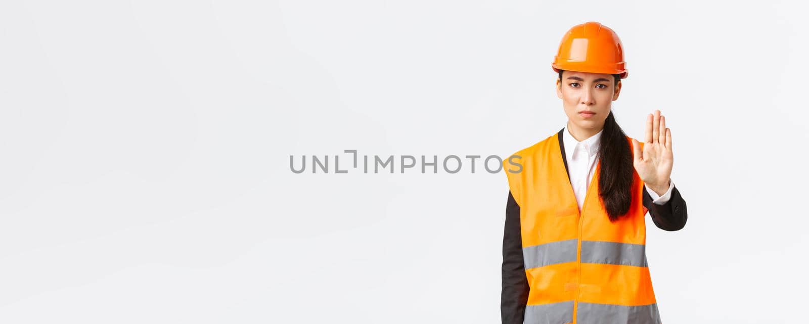Serious-looking disappointed asian female architect, construction manager at working area wearing safety helmet, showing stop gesture, prohibit action, forbid trespassing, white background by Benzoix