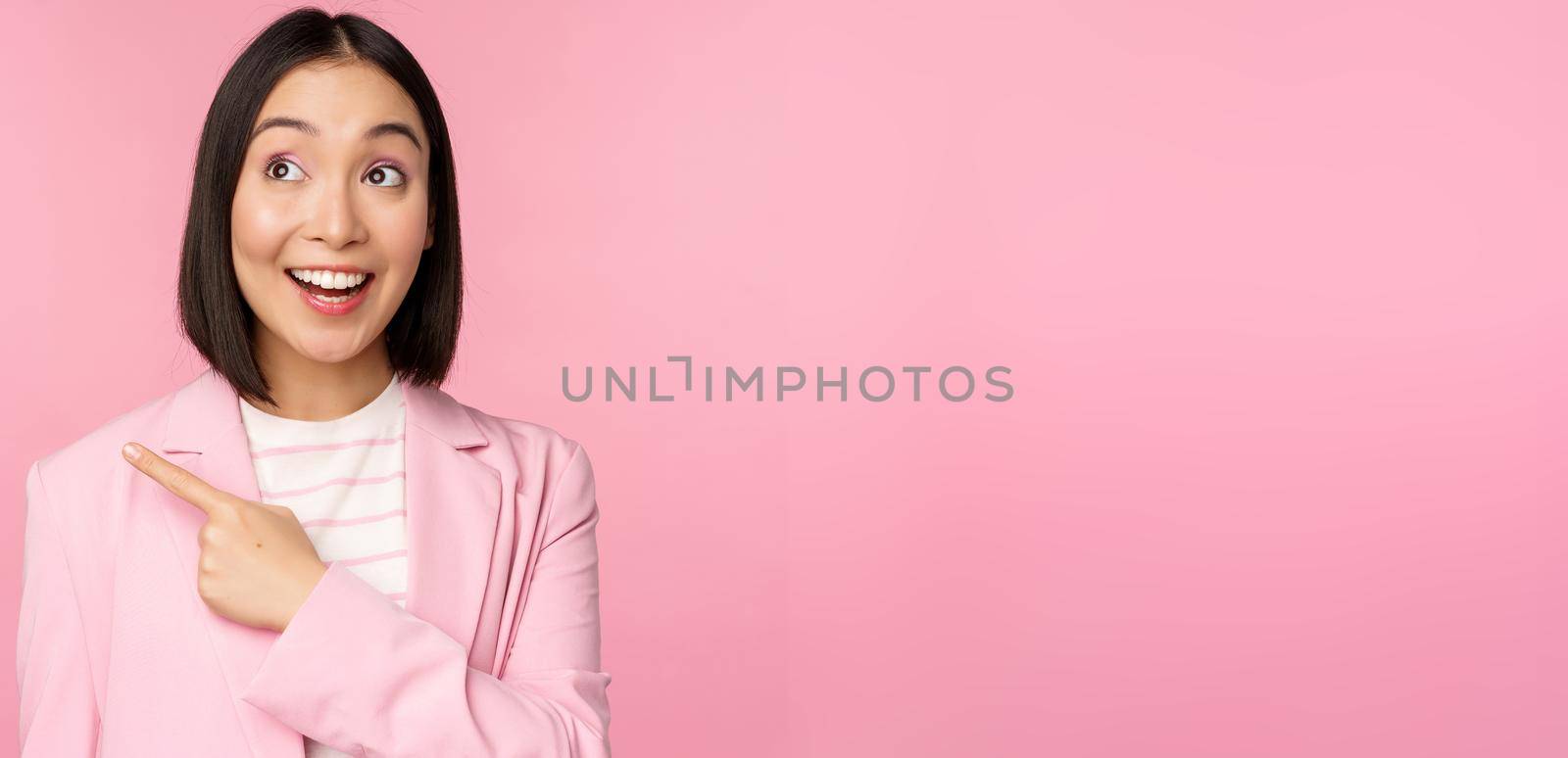 Enthusiastic young asian businesswoman, office employee pointing finger left, looking at banner, advertisement with happy smile, showing advertisement, pink background.