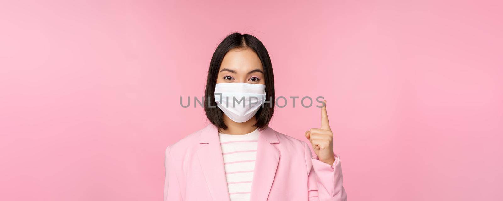 Close up portrait of asian businesswoman in medical face mask and suit, pointing finger up, showing advertisement, top banner, standing over pink background by Benzoix