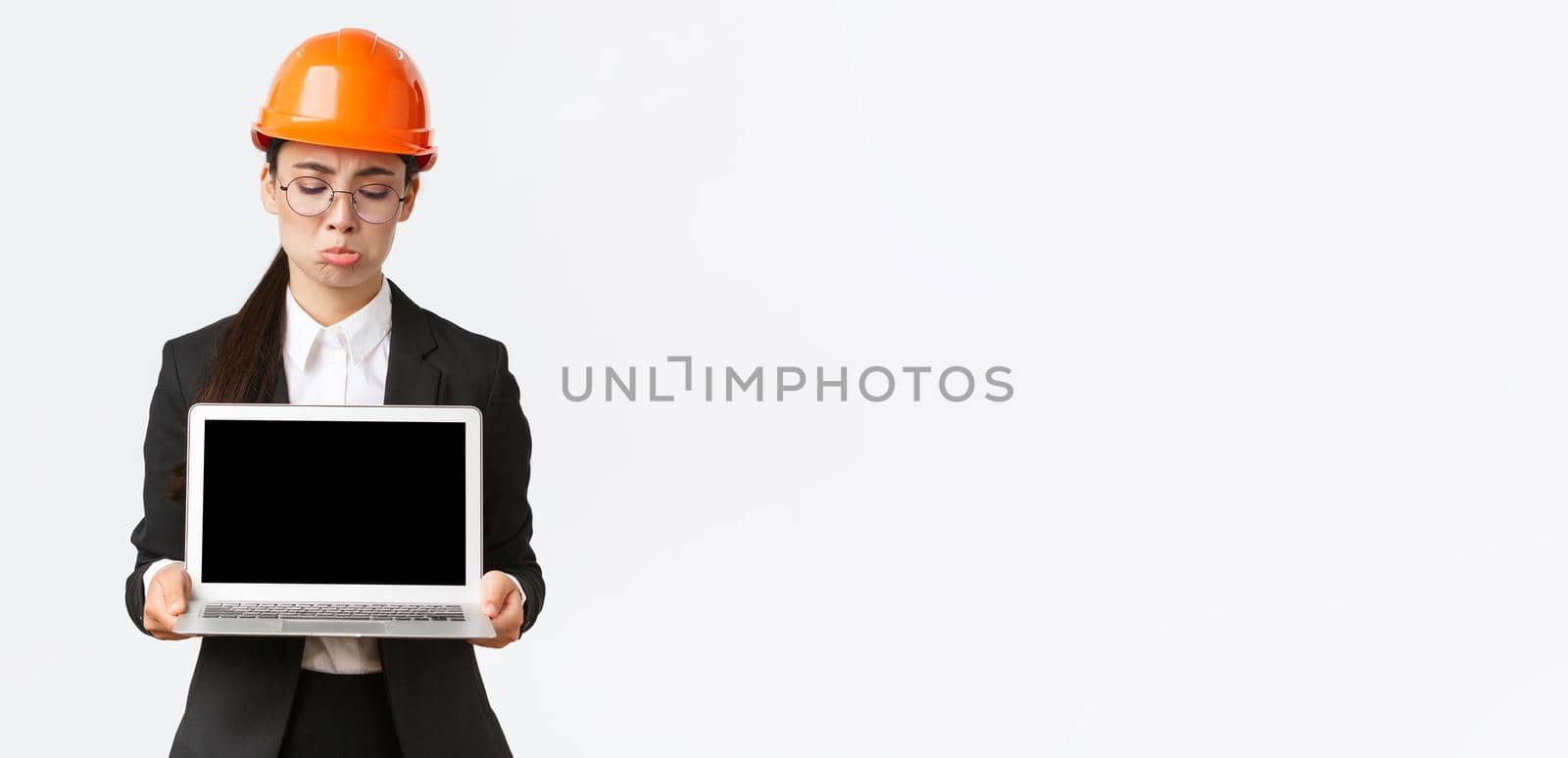 Disappointed gloomy asian female engineer showing bad results, wearing safety helmet and business suit, holding laptop computer and looking at screen displeased, standing white background pouting.