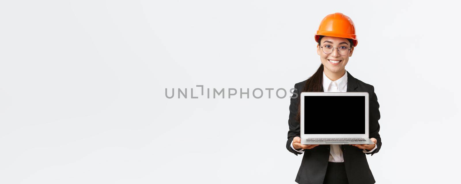 Smiling professional female asian engineer introduce construction plan to investors or clients, standing in safety helmet and suit showing laptop screen with pleased smile, white background by Benzoix