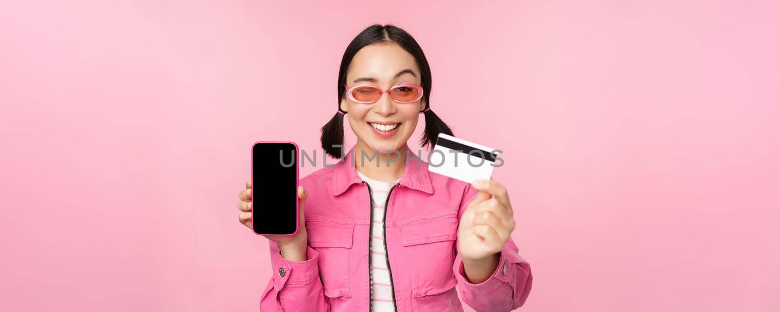 Image of smiling korean woman showing credit card and mobile phone screen, smartphone application interface, paying online, shopping contactless, standing over pink background by Benzoix