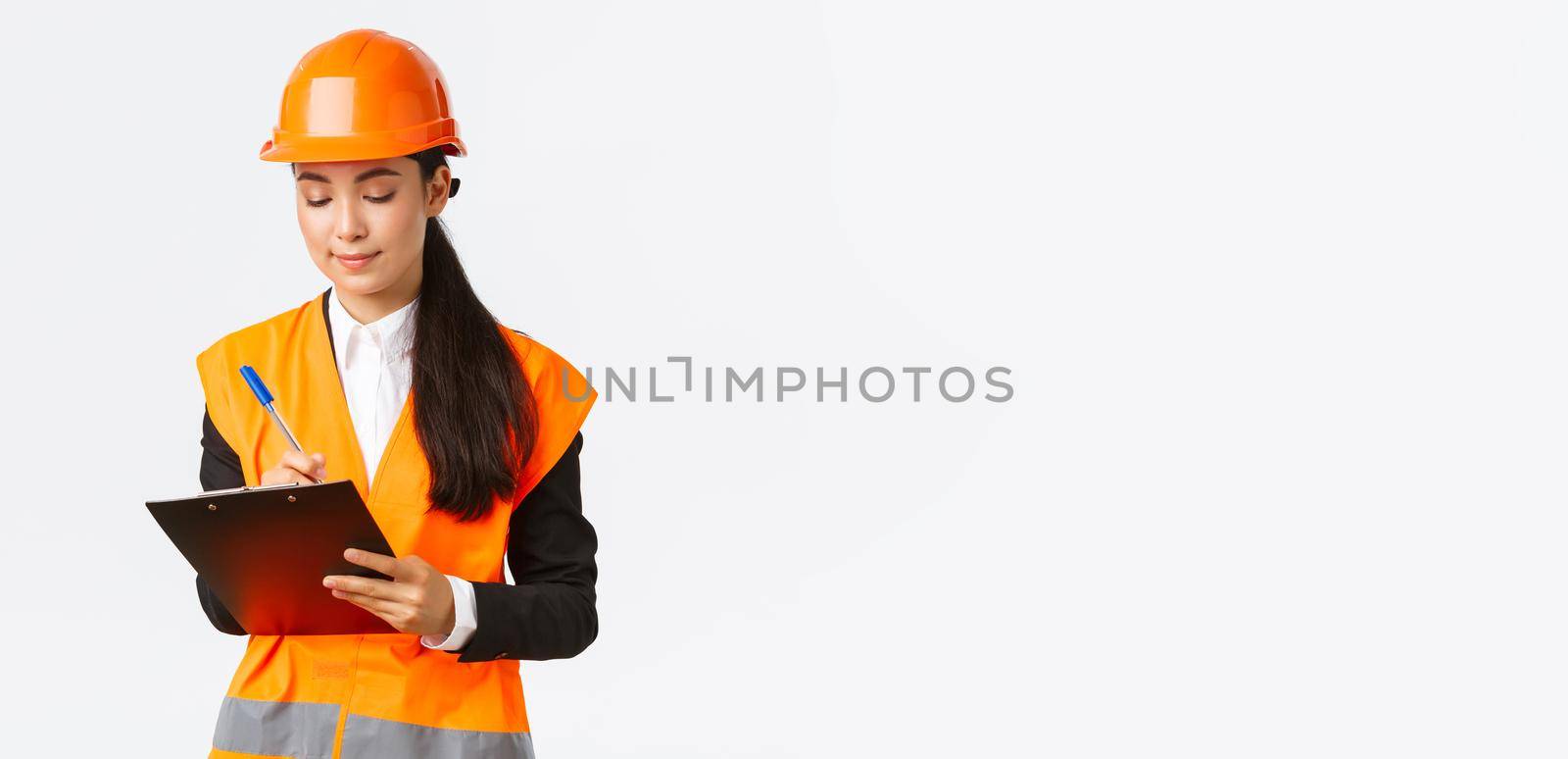 Pleased asian female construction engineer, architect taking notes in clipboard, writing down something during inspection at building area, wearing safety helmet, businesswoman inspect workers.