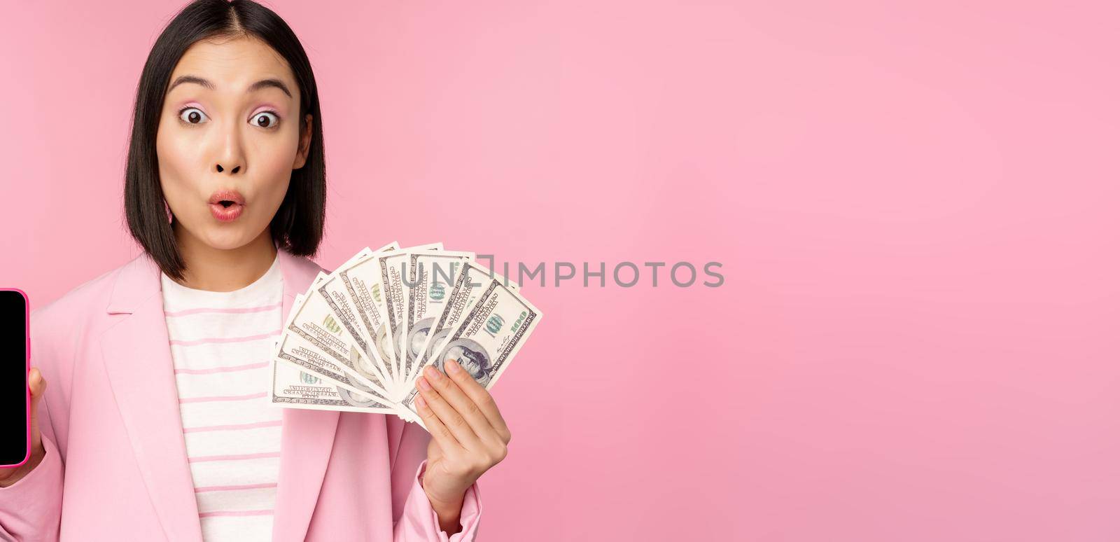 Image of korean successful corporate woman showing money, dollars and smartphone app screen, interface of mobile phone application, concept of investment and finance, pink background.