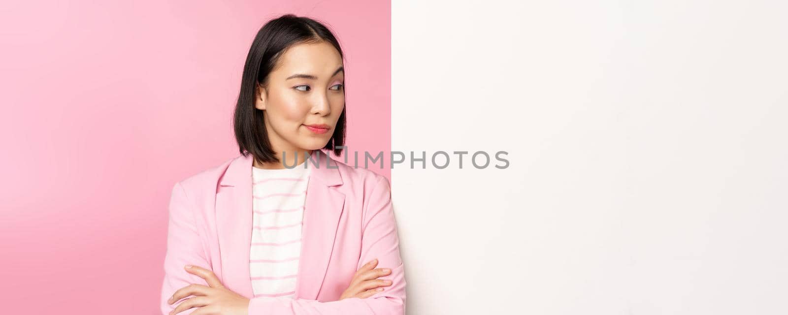 Image of young asian successful businesswoman, corporate lady in suit, standing near white wall info, advertisement on board, cross arms and smiling as professional, pink background.