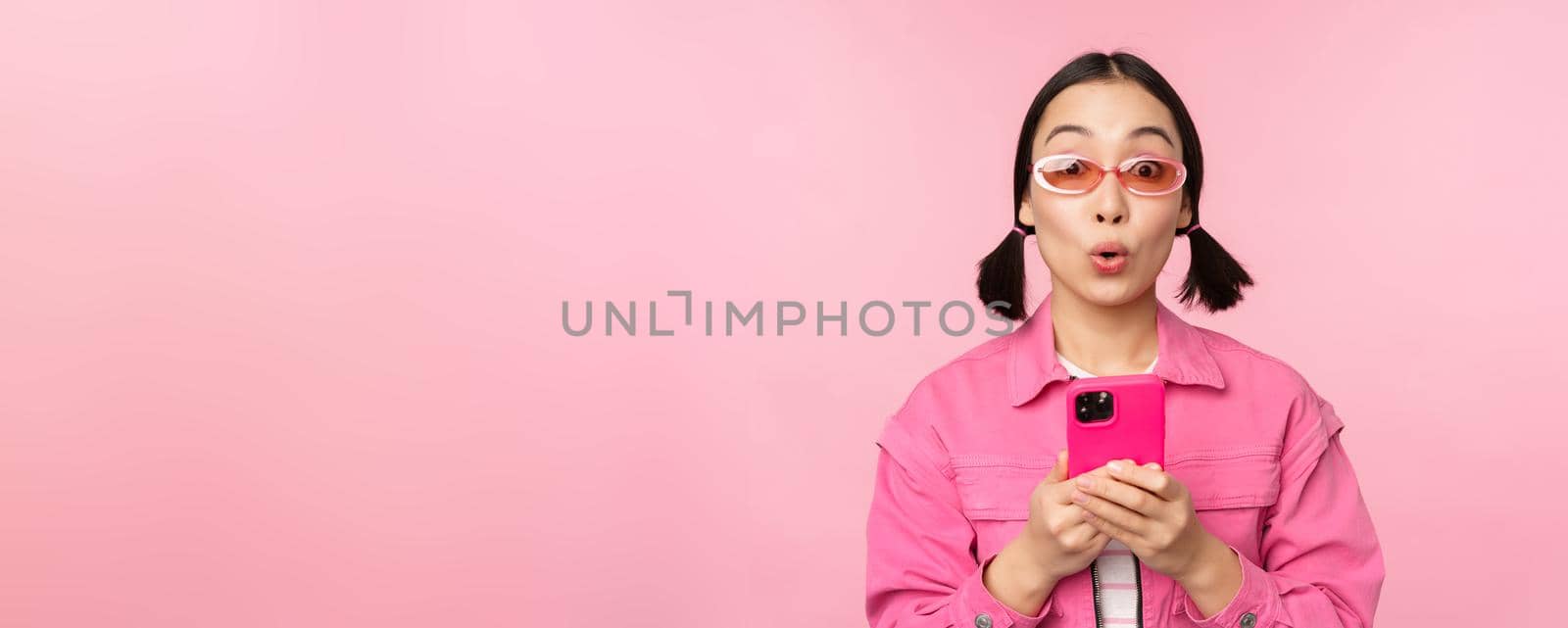 Portrait of korean girl in stylish sunglasses holding mobile phone, using smartphone app, standing over pink background by Benzoix