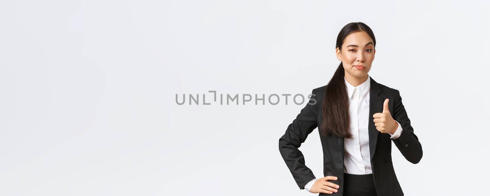 Pleased female entrepreneur, lady boss in black suit satisfied with your work, showing thumbs-up and nod in approval, praise great job, well done, standing white background.