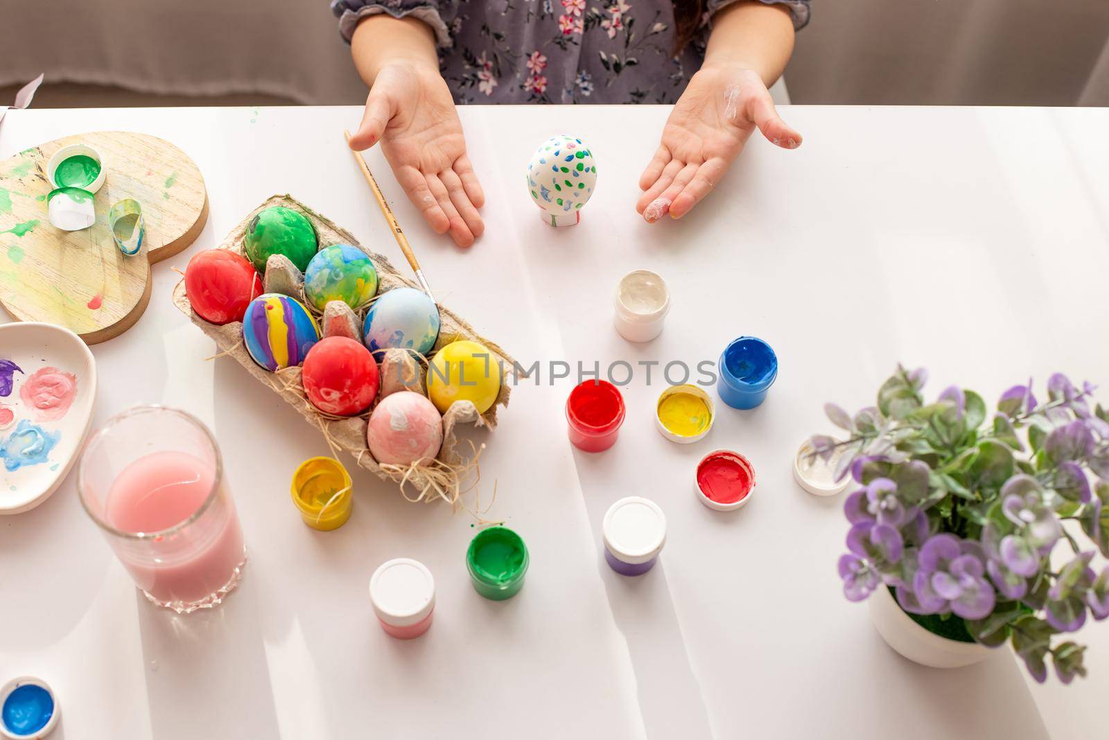 The hands of a little girl, palms up, demonstrate a painted egg, on a white table with multi-colored eggs in a tray, brushes and paints. Top view