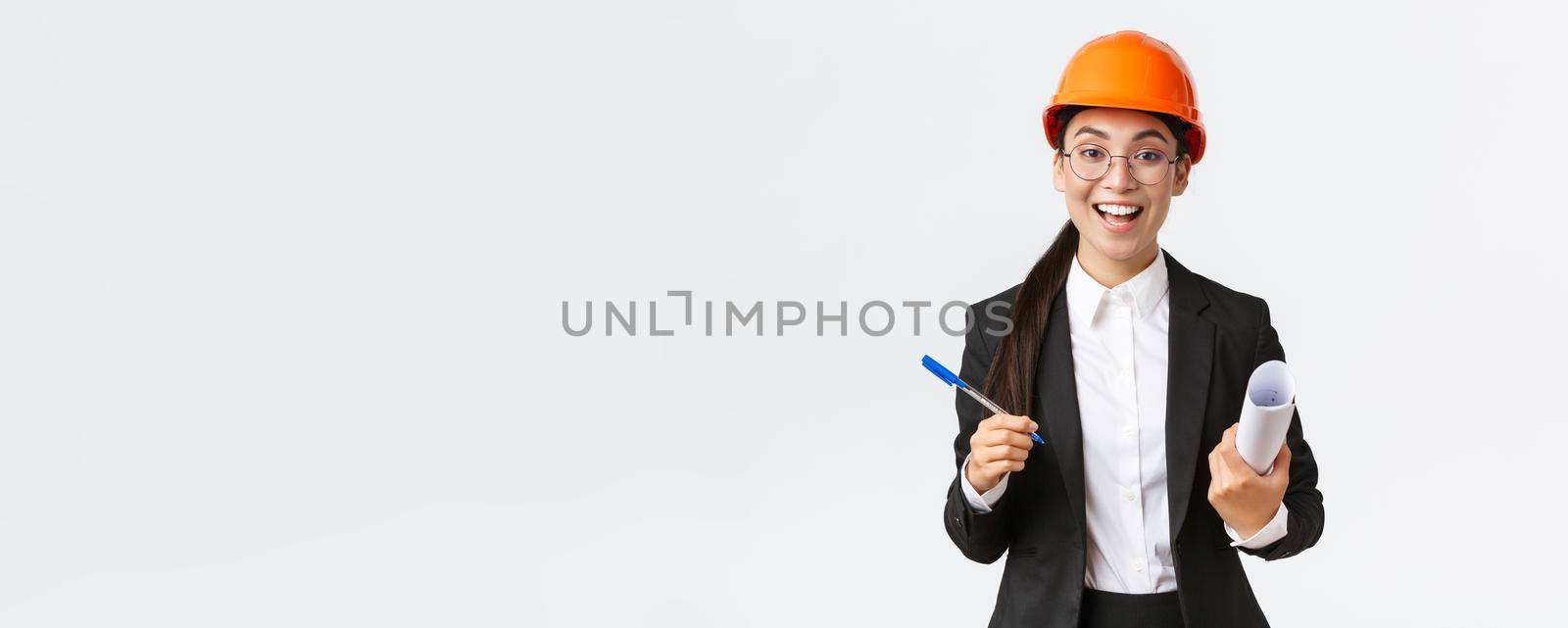 Smiling professional female architect, asian engineer in helmet and business suit, holding blueprints and pen, siging contract, making deal with investors at construction area, white background.