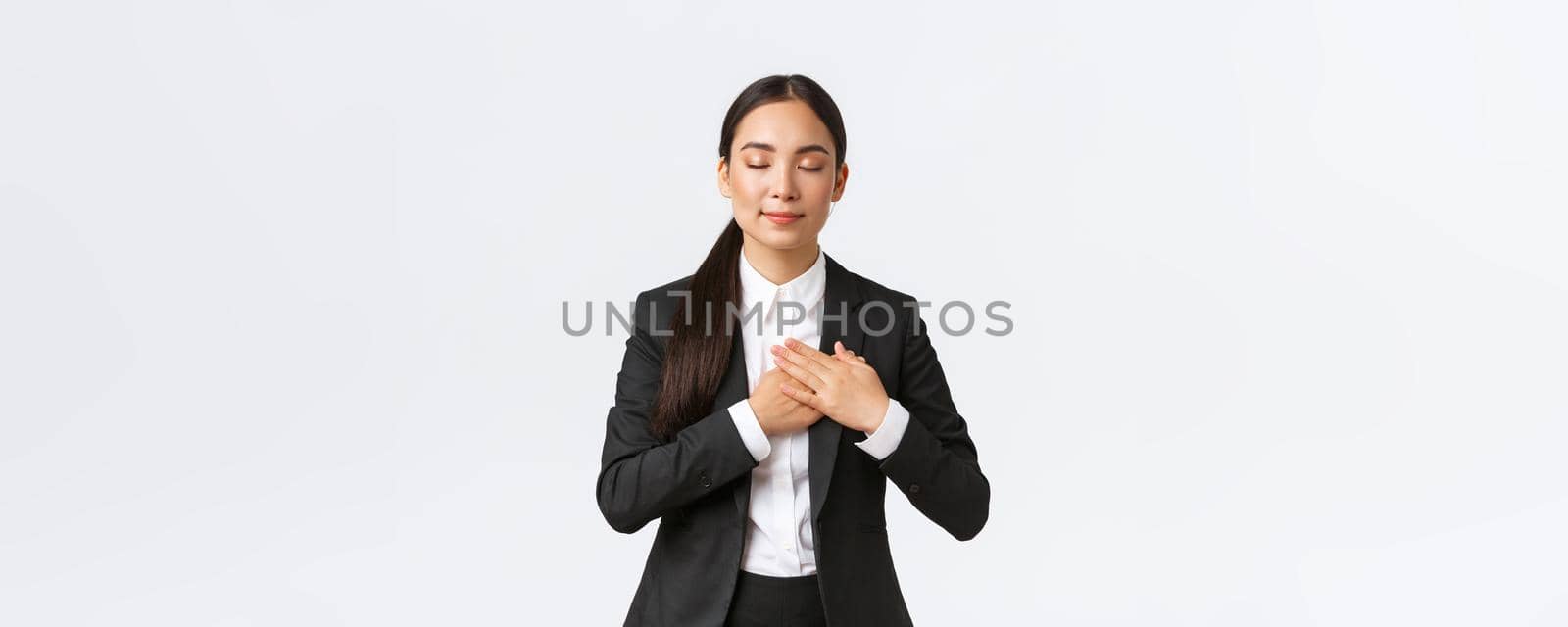 Happy dreamy asian female entrepreneur in suit close eyes and holding hands on heart, remember lovely memory, thinking about something pleasant over white background.