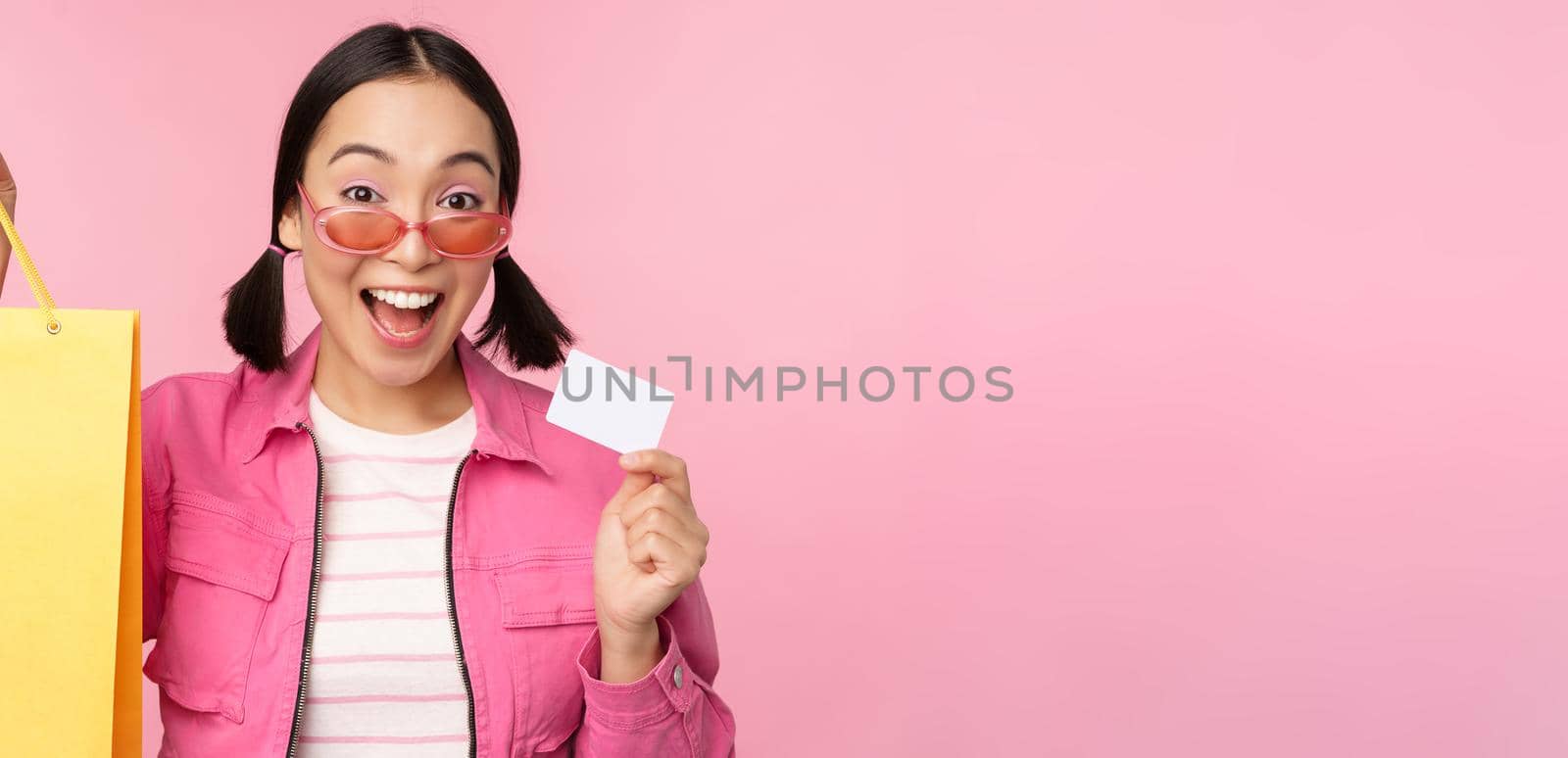 Happy young asian woman shows credit card and shopping bag, store sale announcement, buying smth in shop, posing against pink background.