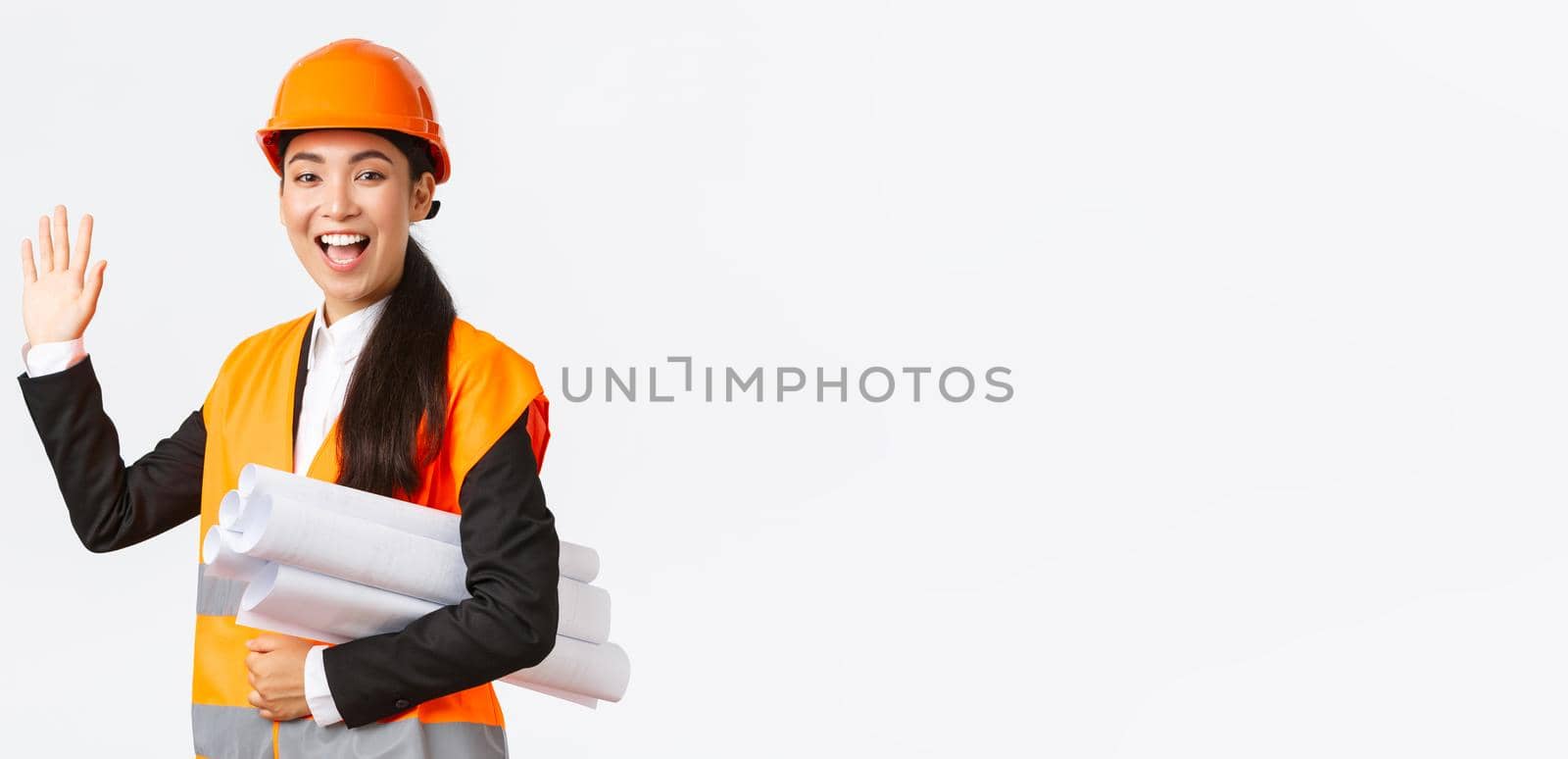 Friendly cheerful asian female construction manager, lead architect in safety helmet and jacket, carry blueprints, documents of building project, waving hand to say hello, greeting someone by Benzoix