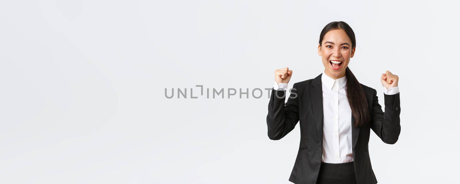 Successful winning female entrepreneur in black suit, fist pump and shouting yes excited, celebrating victory. Businesswoman triumphing over big achievement over white background.