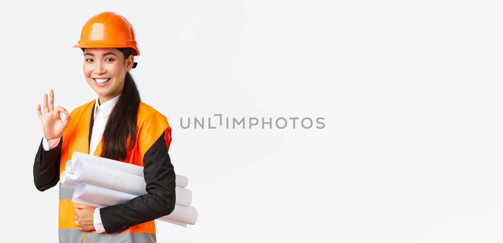 Confident successful female asian construction manager, architect in safety helmet and jacket, showing okay gesture and carry blueprints of building project, guarantee quality, white background.