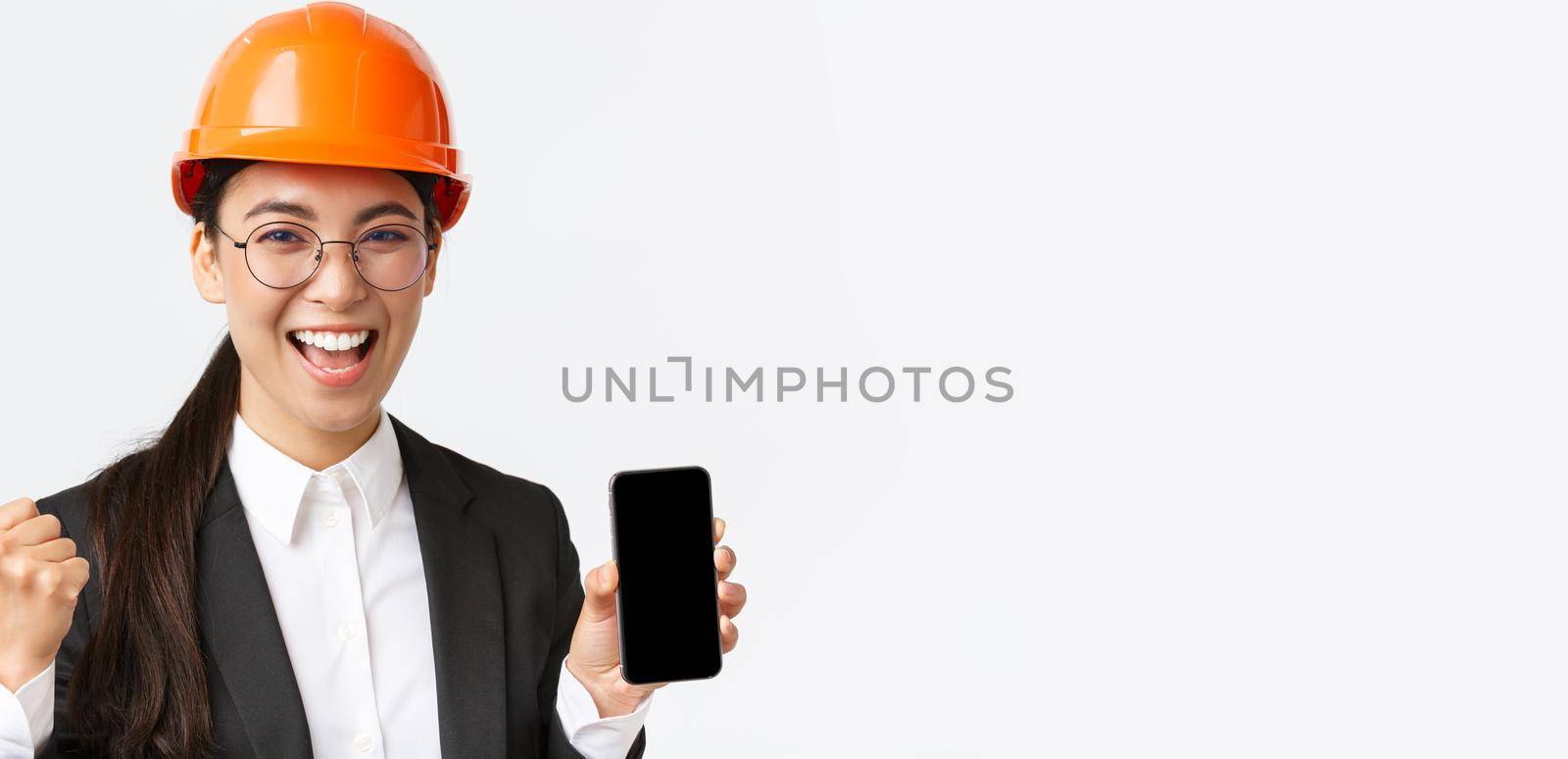 Successful winning asian female engineer, architect in safety helmet and suit, showing smartphone screen, fist pump and shouting yes from rejoice, triumphing over achievement, white background by Benzoix