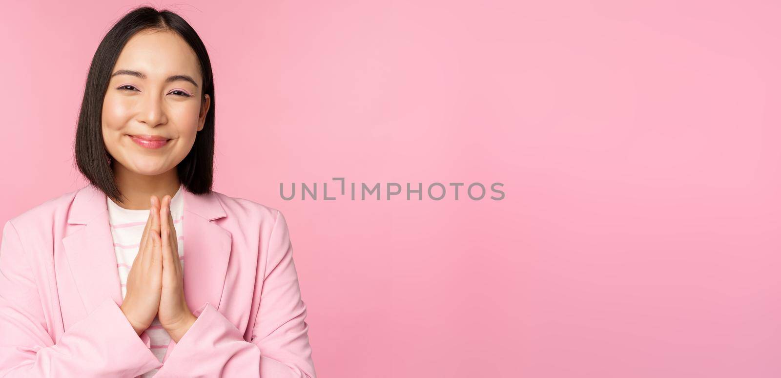 Thank you. Smiling asian saleswoman, corporate lady in suit thanking, holding hands in bag, gratitude gesture, standing over pink background by Benzoix