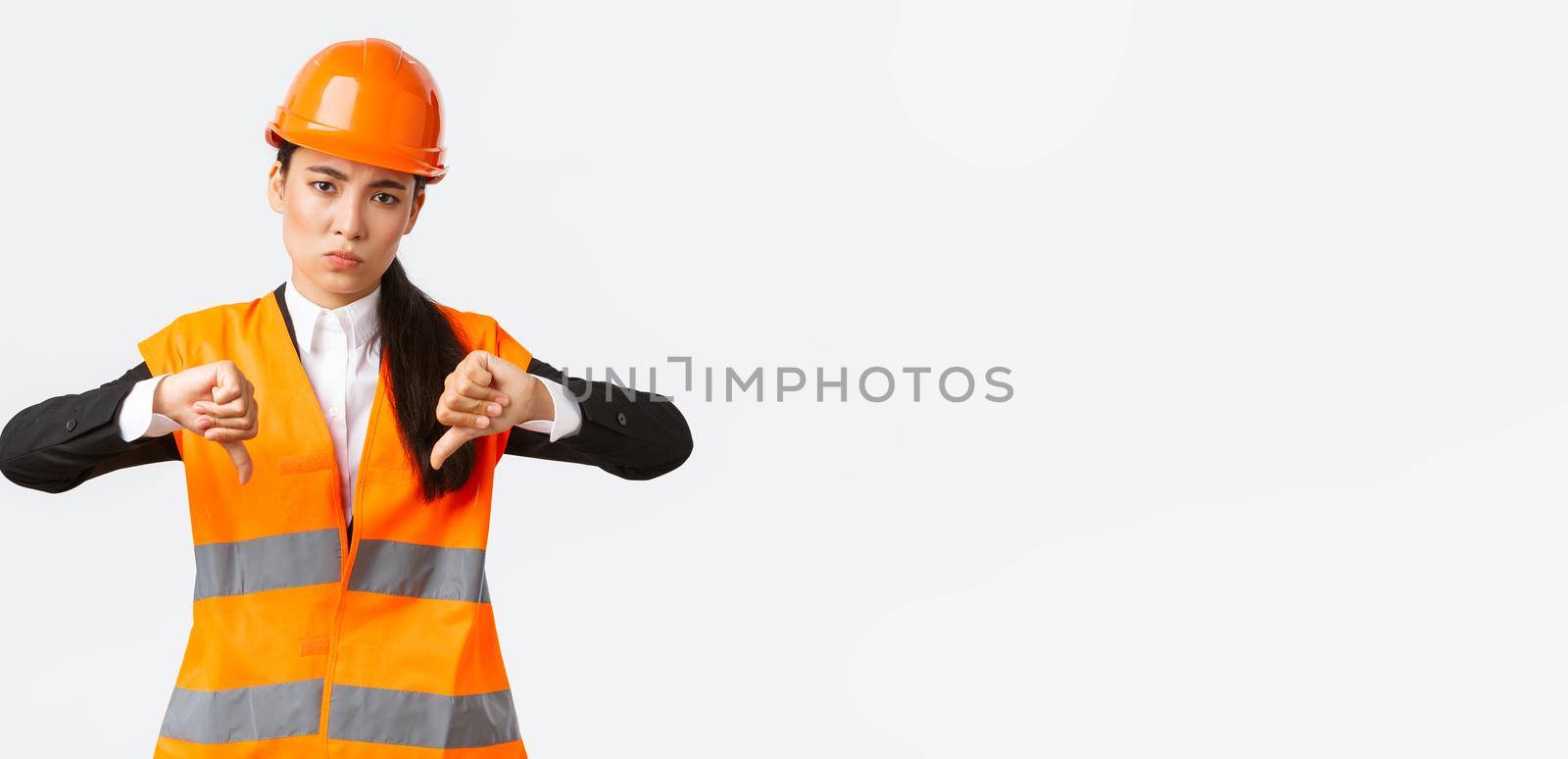 Disappointed and upset asian female chief engineer inspect bad work, showing thumbs-down disappointed, wear safety helmet and complaining weak awful work of architect, white background.