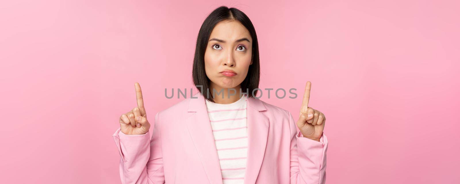 Upset asian corporate woman in office suit, pointing and looking up with disappointed face expression, standing over pink background.