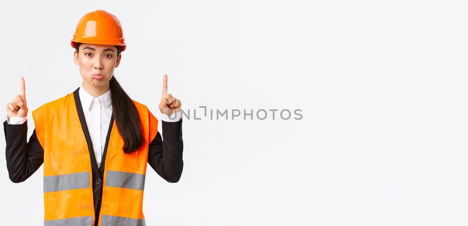 Building, construction and industrial concept. Displeased sad asian female architect complaining, wearing safety helmet and reflective clothing, pouting upset as pointing fingers up.