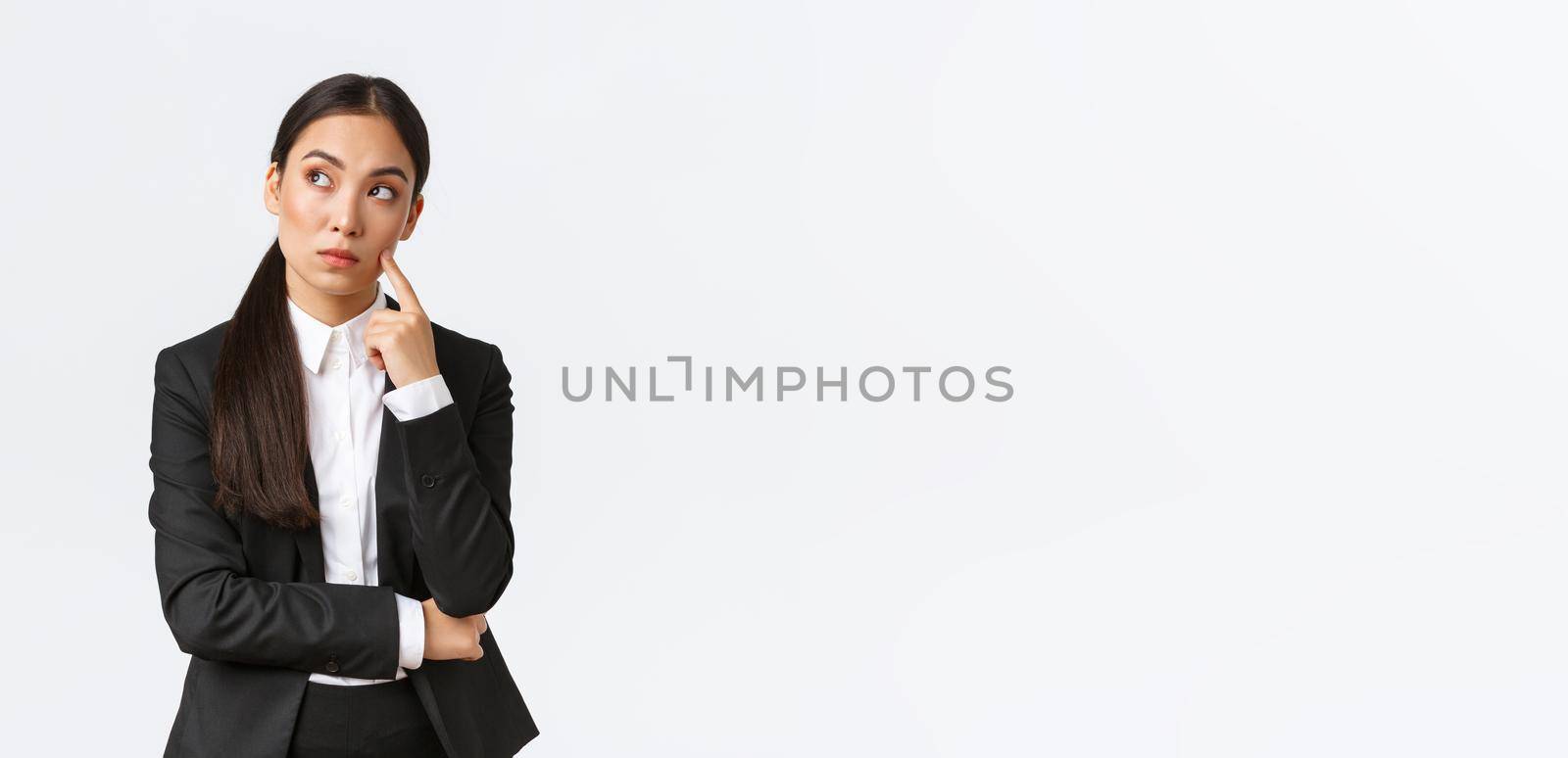 Thoughtful serious-looking asian female manager, office worker in suit thinking about solution, making hard choice, looking away while pondering, standing white background.