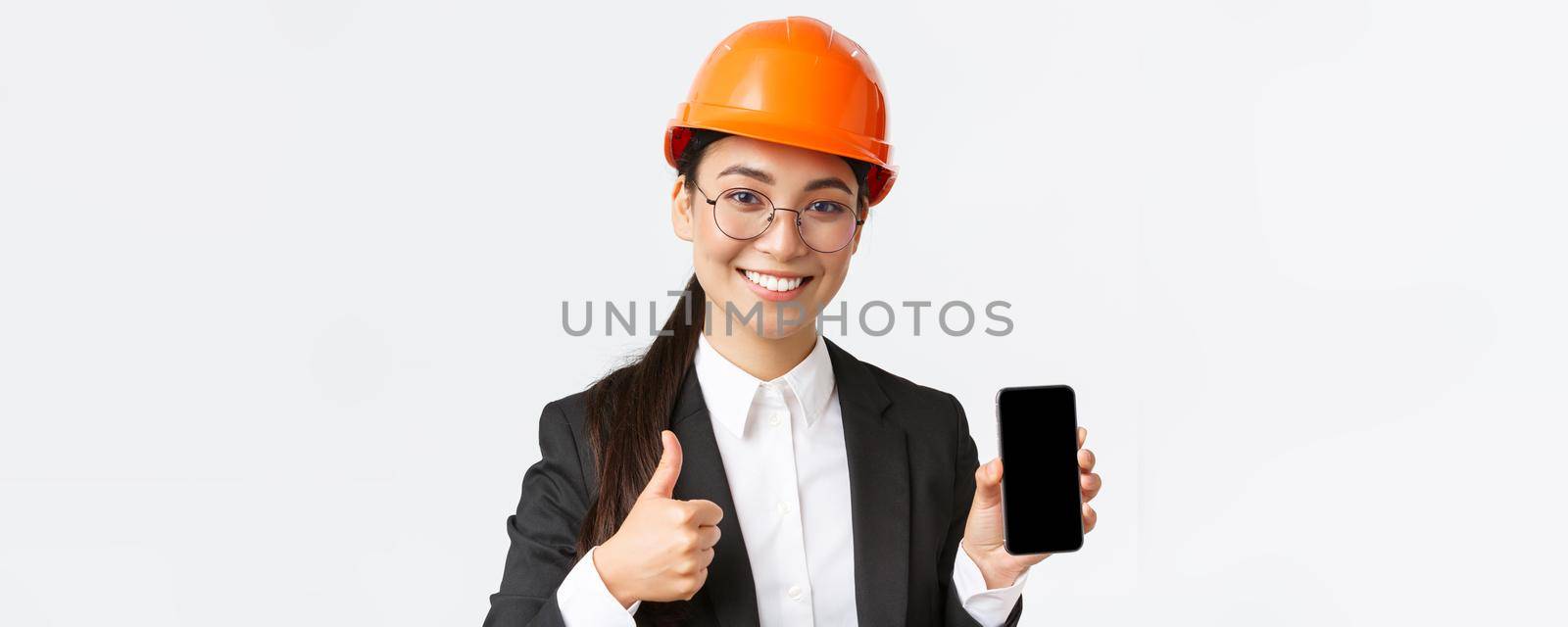 Close-up of professional smiling female engineer, construction manager in business suit and safety helmet, showing smartphone screen and thumbs-up in approval, white background by Benzoix