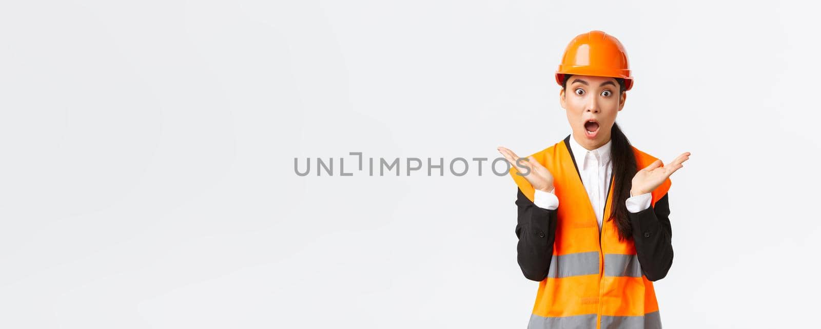 Surprised and shocked asian female engineer in reflective jacket, safety helmet, react to unexpected news, raising hands up and clap impressed, visit construction area, pleased with great work.