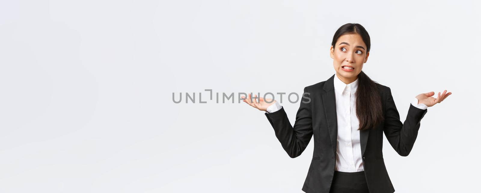 Troubled young female office assistant, saleswoman in black suit shrugging and looking away worried, dont know what do, being confused and unaware, standing nervous over white background.