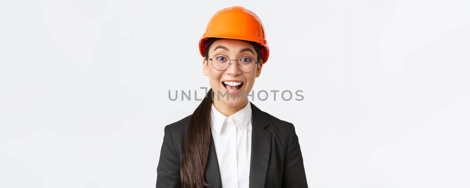 Close-up of excited upbeat asian female chief construction engineer, architect in suit and safety helmet smiling amazed, looking with hope and satisfaction at good work done, white background.