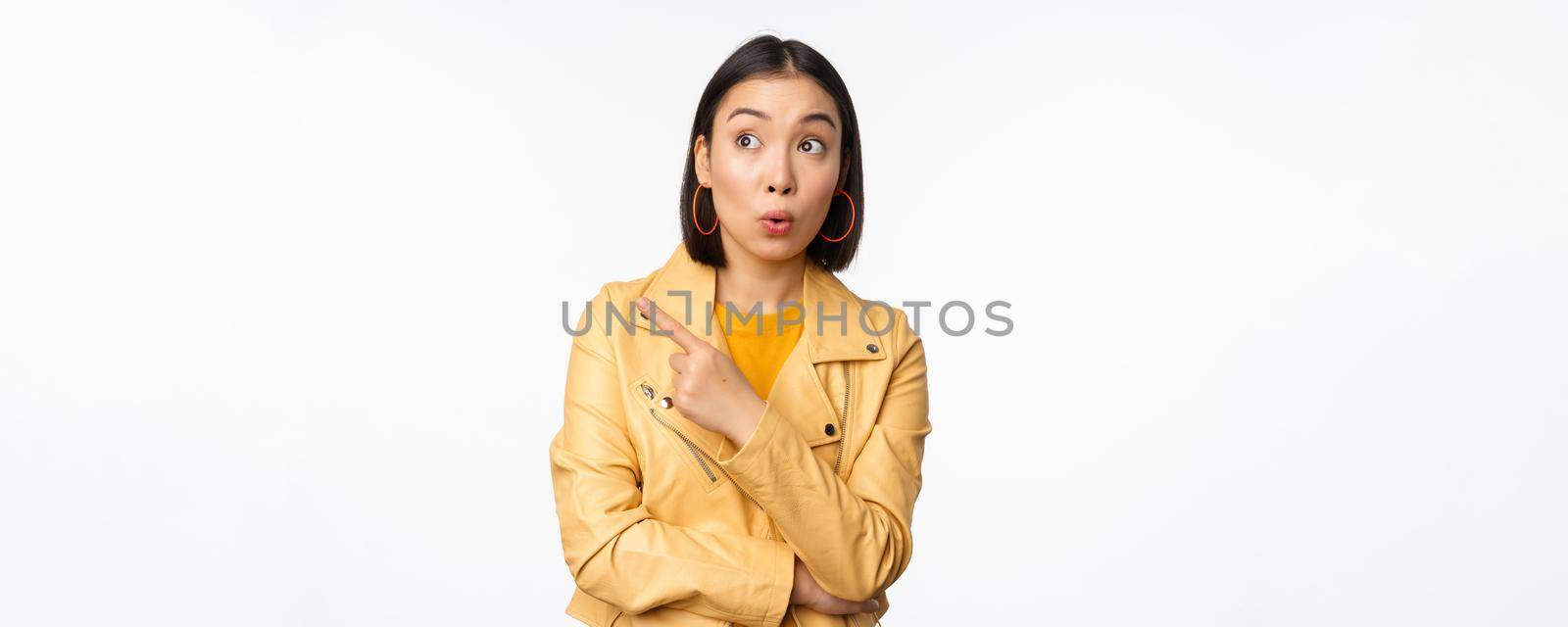 Image of surprised aisan woman pointing finger left, looking at logo or banner with intrigued face expression, standing over white background by Benzoix