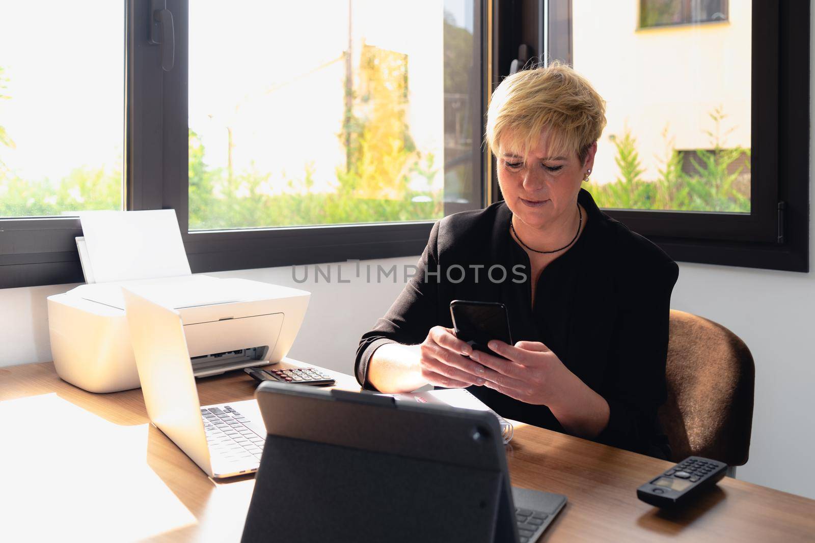 Portrait of a mature, smiling, professional beautician with blonde hair, working in her home office. Small business owner Relaxed atmosphere and soft lighting from window, natural light, work table, desk, computer. Horizontal.