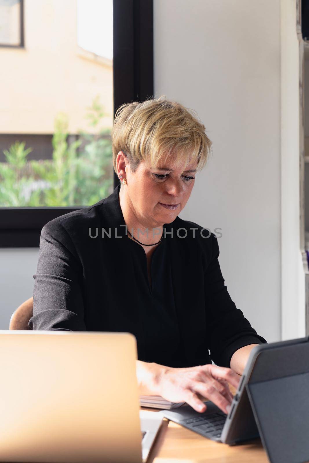 Mature business woman with blonde hair, working in her home office talking on the phone while consulting her laptop. by CatPhotography