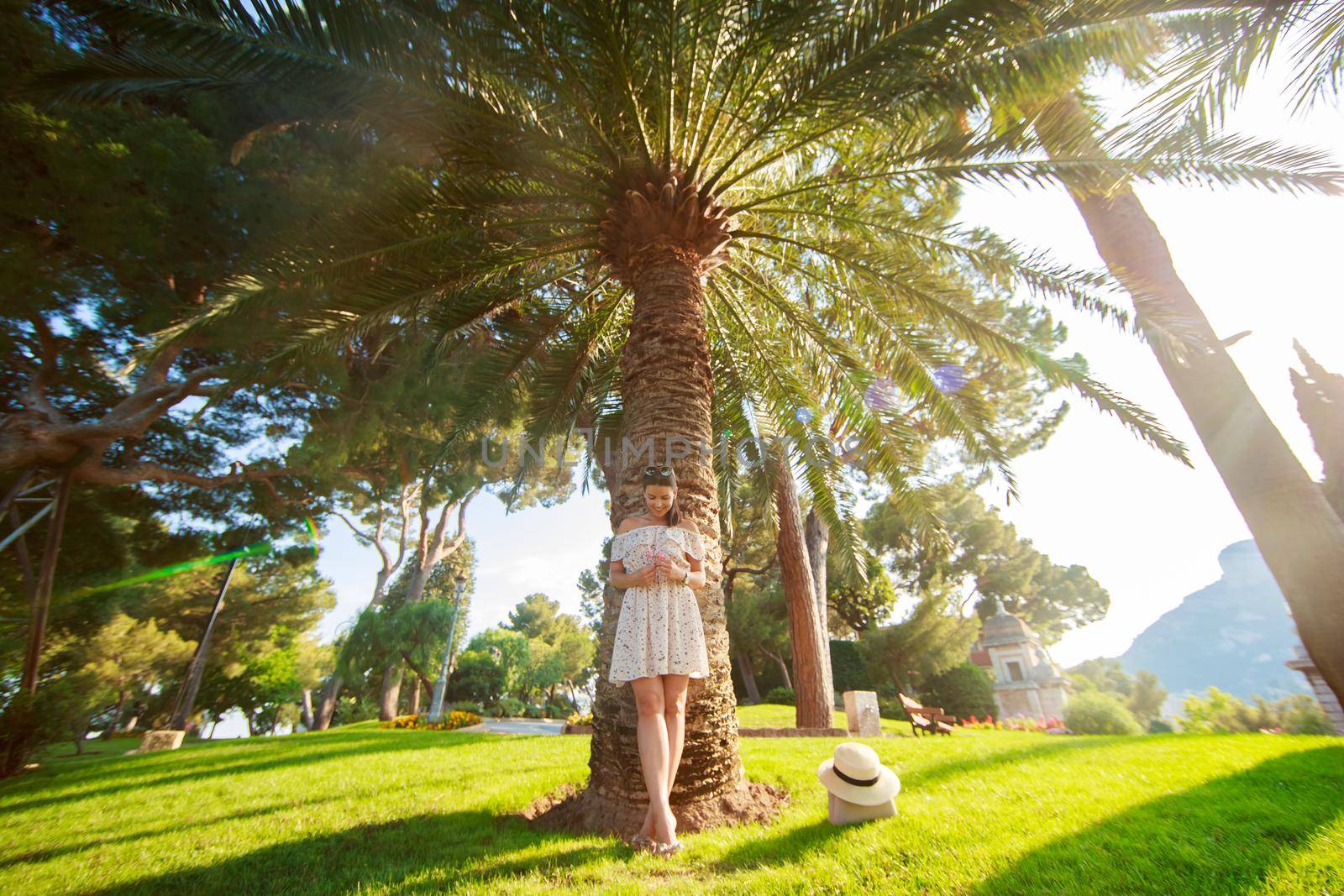The bright beautiful girl in a light dress and hat standing under a palm tree of Monaco in sunny weather in summer. High quality photo
