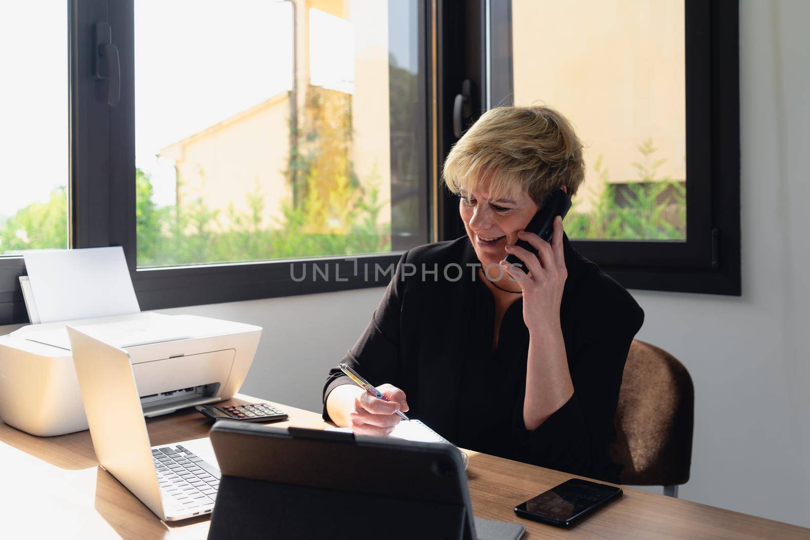 Mature business woman with blonde hair, working in her home office talking on the phone while consulting her laptop. by CatPhotography