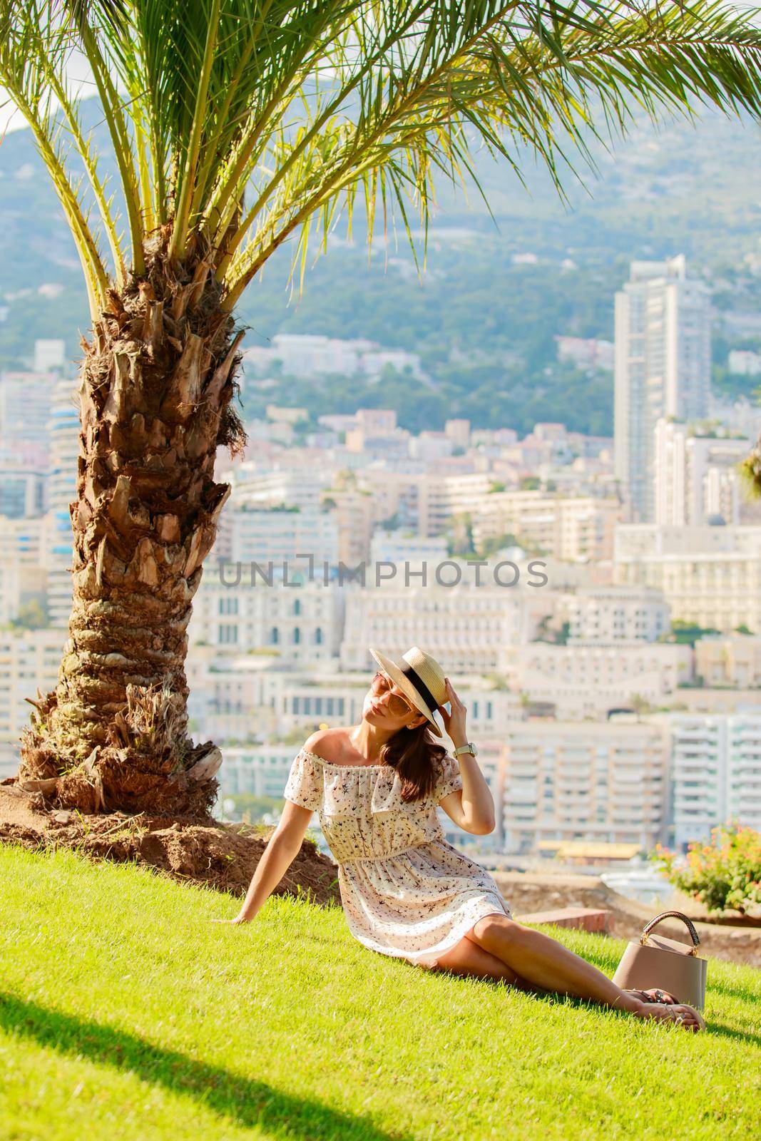 a bright beautiful girl in a light dress and hat sits on the grass under a palm tree in Monaco in sunny weather in summer, streets of old town of Monaco. High quality photo