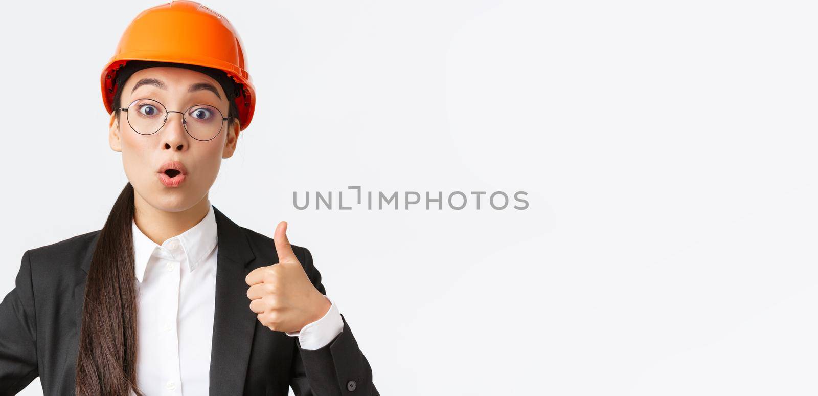 Close-up of professional pleased asian female chief engineer, architect in business suit and safety helmet showing thumbs-up in approval, give permission, recommend construction company.