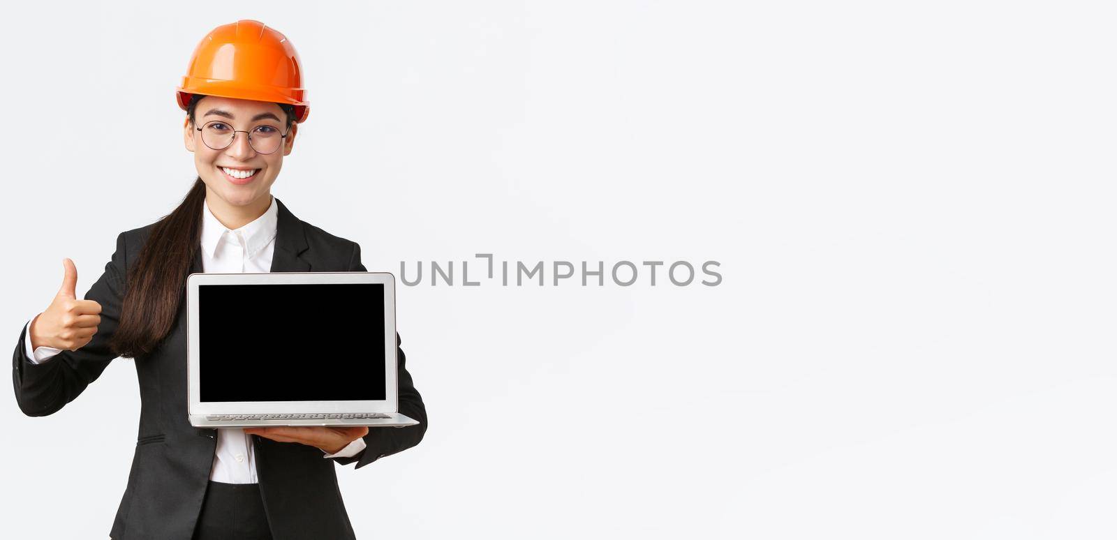 Smiling professional female architect, construction manager at factory showing graph, positive diagram, make thumbs-up in approval and holding laptop facing screen, wear safety helmet by Benzoix