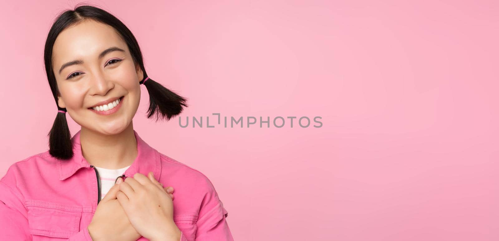 Cute korean girl with silly hairbuns, looking happy and grateful, thank you pose, holding hands on heart flattered, posing against pink background.