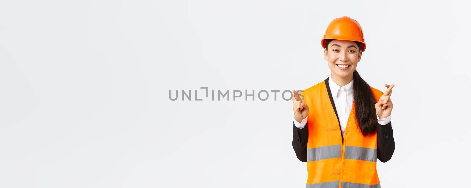 Hopeful and optimistic smiling asian female engineer, construction manager in safety helmet having faith in project, cross fingers good luck and looking positive, standing white background.