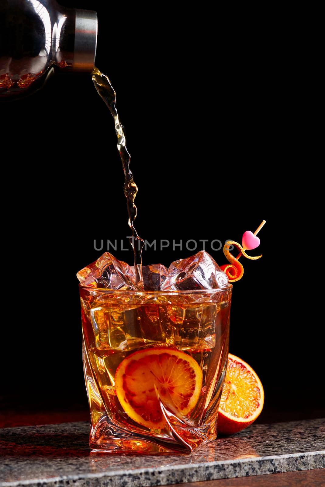 Alcohol cocktail pouring from shaker into gless with orange peel ice on stone desk. by PhotoTime