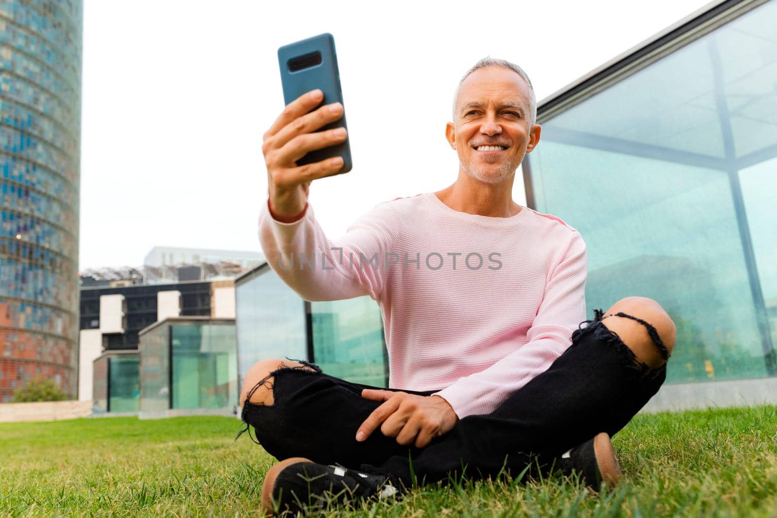 Caucasian man takes selfie using mobile phone sitting on the grass in city park. by Hoverstock