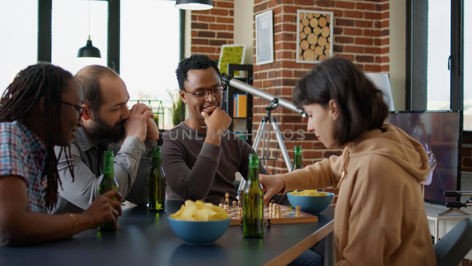 Diverse group of friends playing strategic board games at leisure gathering with beer bottles and snacks. People laughing and enjoying meeting to play chess match together for entertainment.